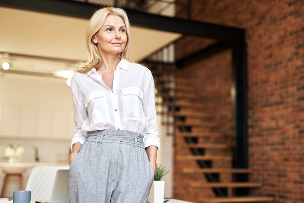 Succesvolle rijpe blonde zakenvrouw kijkt opzij terwijl ze poseert terwijl ze op kantoor staat