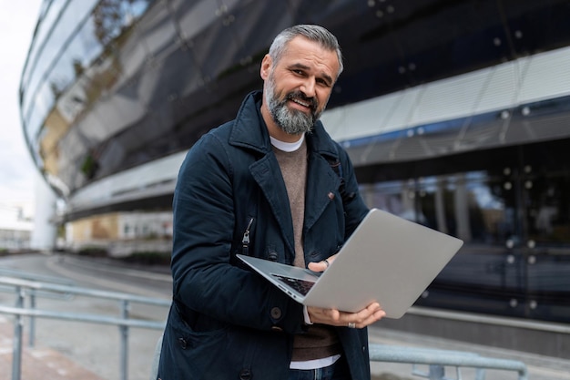 Succesvolle volwassen man met een laptop in zijn handen tegen de achtergrond van een kantoorgebouw buiten