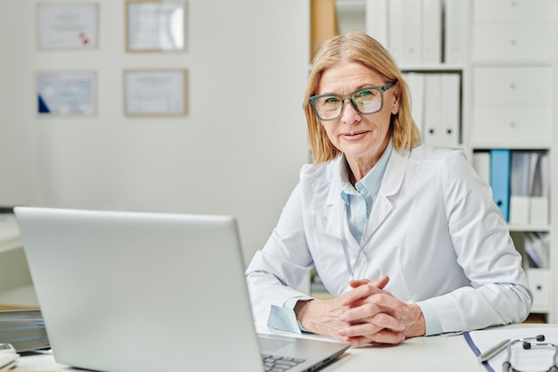 Succesvolle volwassen vrouwelijke clinicus in brillen en whitecoat zittend aan bureau