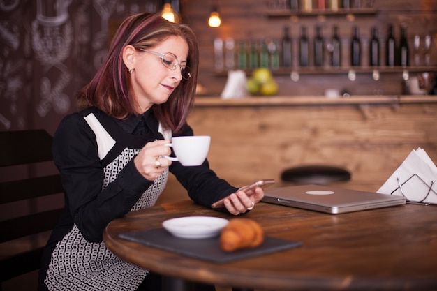 Succesvolle volwassen zakenvrouw die naar haar telefoon kijkt terwijl ze een kopje koffie drinkt in een vintage pub