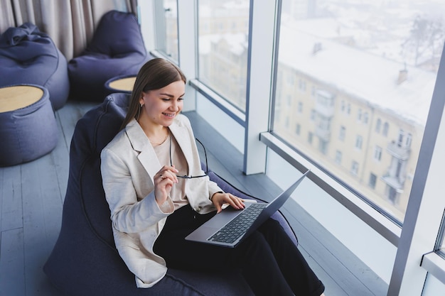 Succesvolle vrouw in vrijetijdskleding typt op een netbook terwijl ze aan een nieuw project werkt terwijl ze aan een bureau zit in een moderne werkruimte Zakelijke dame in een jas en bril Telewerken