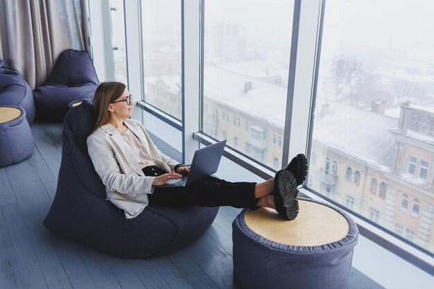 Succesvolle vrouw in vrijetijdskleding typt op een netbook terwijl ze aan een nieuw project werkt terwijl ze aan een bureau zit in een moderne werkruimte Zakelijke dame in een jas en bril Telewerken