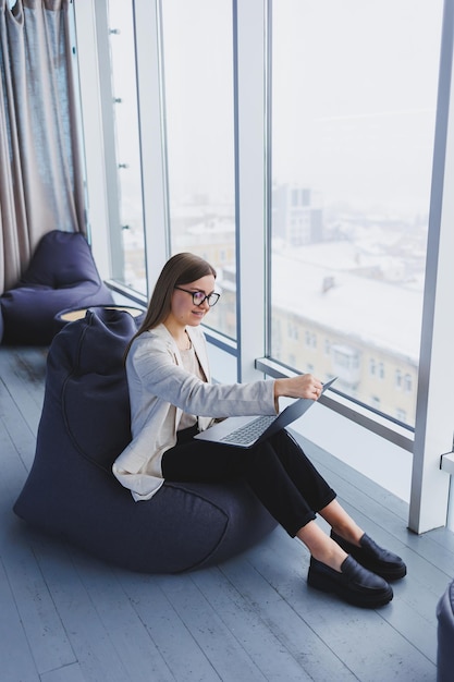 Succesvolle vrouw in vrijetijdskleding typt op een netbook terwijl ze aan een nieuw project werkt terwijl ze aan een bureau zit in een moderne werkruimte Zakelijke dame in een jas en bril Telewerken