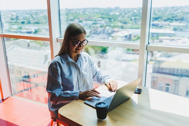 Succesvolle vrouw kijkt naar een laptop in een café en drinkt koffie een jonge lachende vrouw met een bril zit aan een tafel bij het raam met een telefoon freelance en werk op afstand moderne vrouwelijke levensstijl