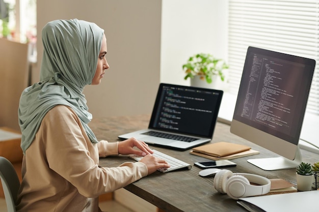 Succesvolle vrouw uit het Midden-Oosten met een lichtblauwe hijab die aan het bureau zit en aan computercode werkt