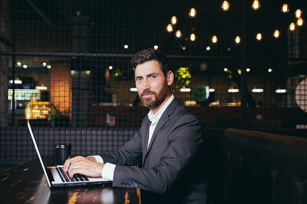 Succesvolle zakenman die op laptop werkt tijdens de lunchpauze die in het restaurant van het hotel zit en serieus naar de camera kijkt met een kopje koffie