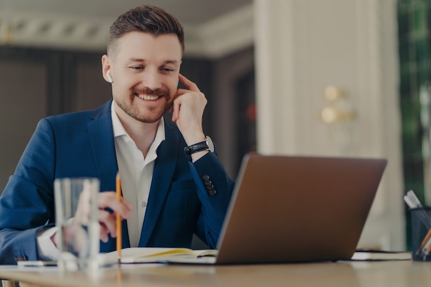 Succesvolle zakenman in draadloze oortelefoons die webconferentie houdt op zijn kantoor met werknemers, een formeel donkerblauw pak draagt terwijl hij voor een laptop zit met een potlood in de hand, thuis werkend