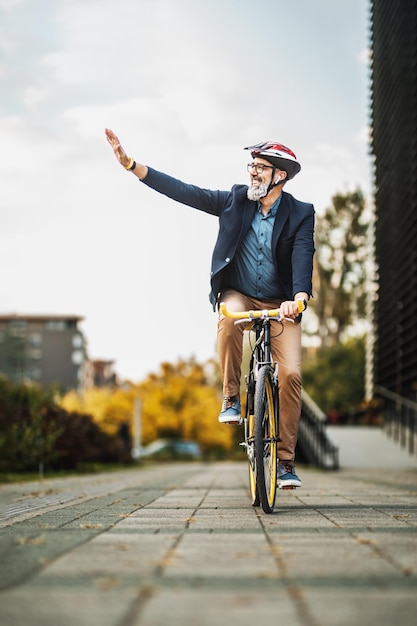 Succesvolle zakenman van middelbare leeftijd die naar iemand zwaait terwijl hij fietst op weg naar zijn werk door de stad.