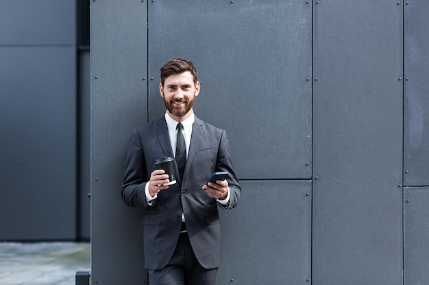 Succesvolle zakenmanmakelaar in een pak aan de telefoon in de buurt van het kantoor buiten, bankier met een kopje koffie tijdens de lunchpauze glimlachend