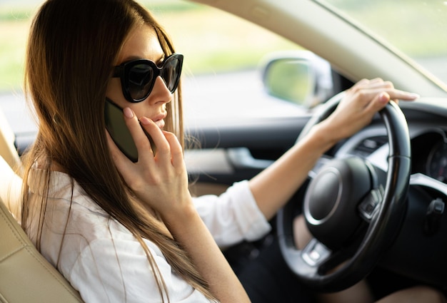 Succesvolle zakenvrouw die een auto bestuurt in een zonnebril met een mobiele telefoon in haar handen