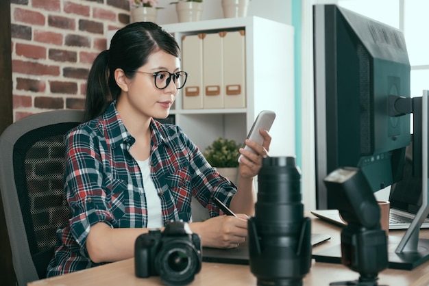 succesvolle zelfverzekerde vrouwelijke fotograaf die op een bureau zit en foto's bewerkt en een mobiele smartphone gebruikt die de foto serieus controleert.