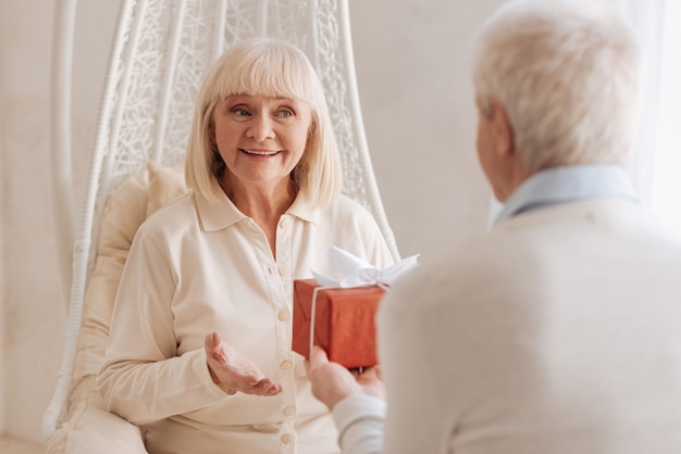 Such a surprise. Cheerful delighted senior woman smiling and looking at her husband while receiving a present from him