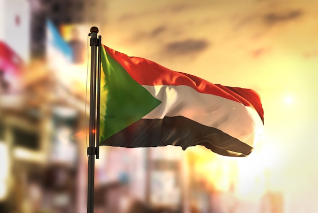 Sudan Flag Against City Blurred Background At Sunrise Backlight