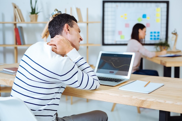 Suffer pain. Frustrated man touching his neck and putting arms on the table while looking at screen of computer