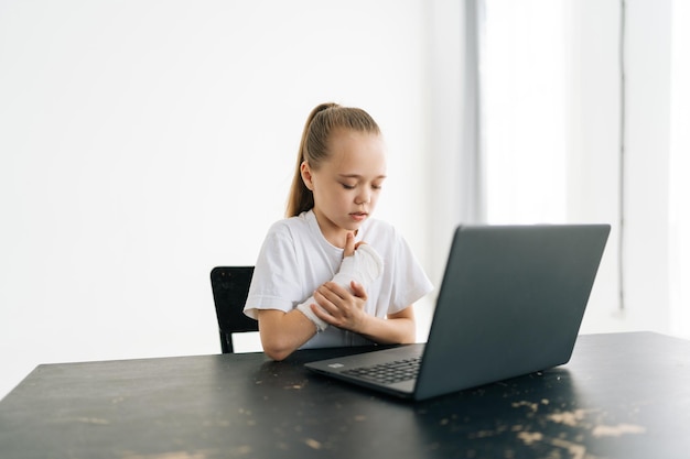 가벼운 방에 노트북 컴퓨터가 있는 탁자에 앉아 있는 다친 팔을 만지는 흰색 석고 붕대에 감긴 손이 부러진 어린 소녀가 고통을 겪고 있습니다.