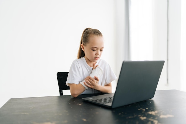 Suffering from pain little girl with broken hand wrapped in white plaster bandage touching injured arm sitting at table with laptop computer in light room