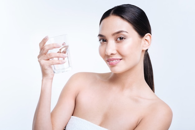 Sufficient water level. Beautiful dark-haired young woman holding a glass of water and smiling  while standing isolated on a white wall