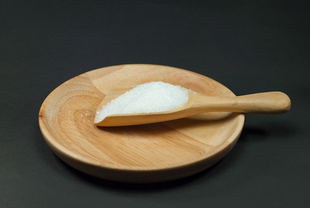 Sugar in a wooden spoon placed on a wooden plate and a black background.