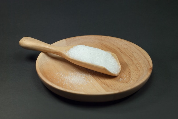 Sugar in a wooden spoon placed on a wooden plate and a black background.