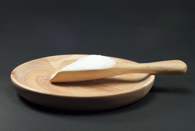 Sugar in a wooden spoon placed on a wooden plate and a black background.