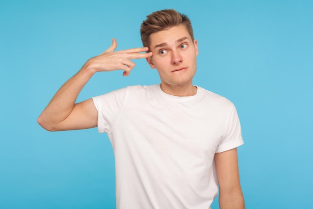 Suicide gesture Portrait of depressed unhappy man in tshirt pointing finger gun to head and looking with tedious indifferent expression bored of life indoor studio shot isolated on blue background