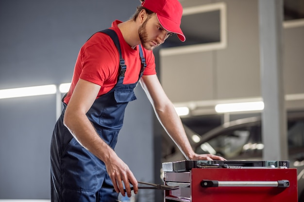 Suitable option. Serious interested young man in red tshirt and blue overalls picking suitable piece near toolbox