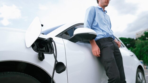 Suitclad businessman with progressive ambition leaning on his electric vehicle while standing on a charging station with a power cable plug and a renewable energypowered electric vehicle