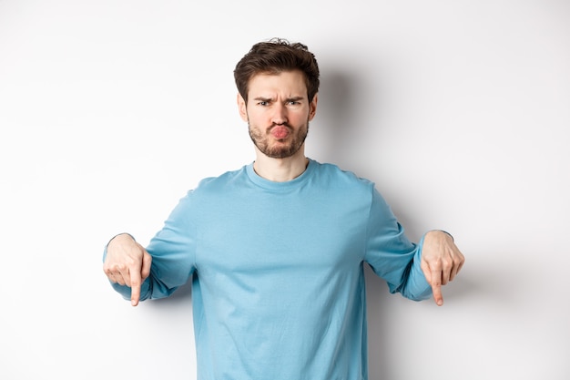 Sulking man pointing fingers down at something bad, frowning and complaining, standing over white background.