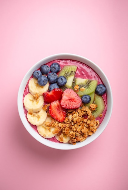 Summer acai smoothie bowl with strawberries, banana, blueberries, kiwi fruit and granola on pastel pink background. Breakfast bowl with fruit and cereal, close-up, top view, healthy food