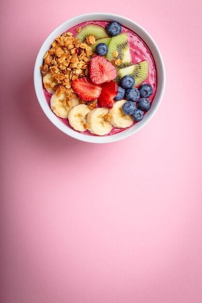 Summer acai smoothie bowl with strawberries, banana, blueberries, kiwi fruit and granola on pastel pink background. Breakfast bowl with fruit and cereal, close-up, top view, space for text