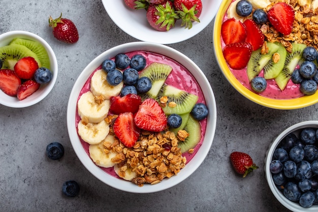 Summer acai smoothie bowls with strawberries, banana, blueberries, kiwi fruit and granola on gray concrete background. Breakfast bowl with fruit and cereal, close-up, top view, healthy food