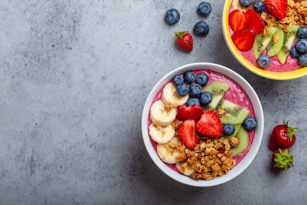 Summer acai smoothie bowls with strawberries, banana, blueberries, kiwi fruit and granola on gray concrete background. Breakfast bowl with fruit and cereal, close-up, top view, space for text