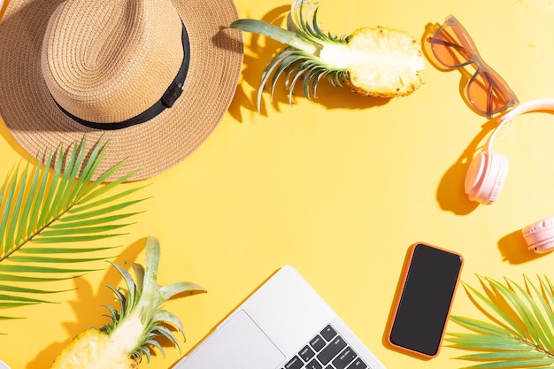 Summer backdrop with sedge hat, laptop, phone and pineapple on a yellow summer background