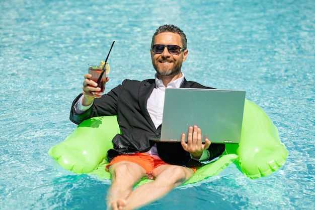 Summer business dreams millennial business man in suit floating with cocktail and laptop in swimming