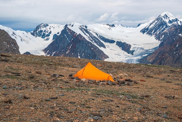 Summer camping in mountain Bright alpine landscape with vivid orange tent at very high altitude with view to high mountain and large glacier in dramatic clouds Awesome mountain scenery with tent