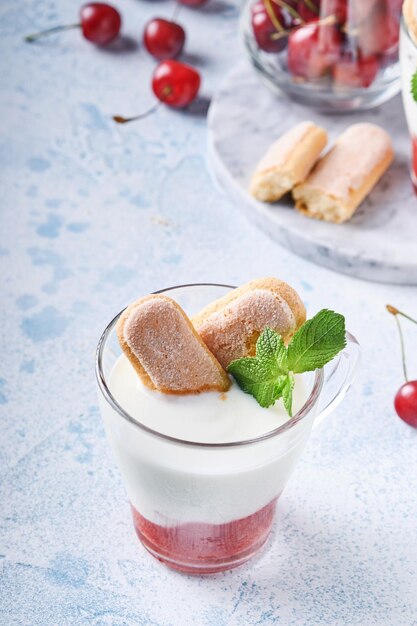 Sfogliatina estiva alle ciliegie con savoiardi e crema di formaggio in vetro su fondo grigio chiaro. torta tiramisù tradizionale con frutti di bosco freschi. messa a fuoco selettiva.