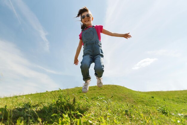 summer, childhood, leisure and people concept - happy little girl jumping high over green field and blue sky outdoors