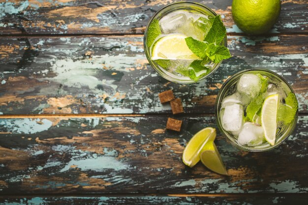 Summer drink in glass and ingredients for making Mojito with ice, brown sugar, lime slices, fresh mint on wooden rustic background, space for text. Cold refreshing cocktail for summer party and fun