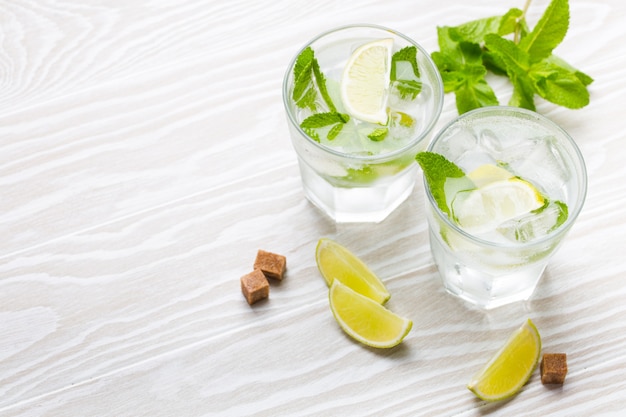 Bevande estive con ghiaccio, zucchero di canna, fette di lime, menta fresca in bicchieri, fondo rustico in legno bianco, spazio per il testo. limonate/mojito rinfrescanti fredde ottime per feste estive e divertimento, copia spazio