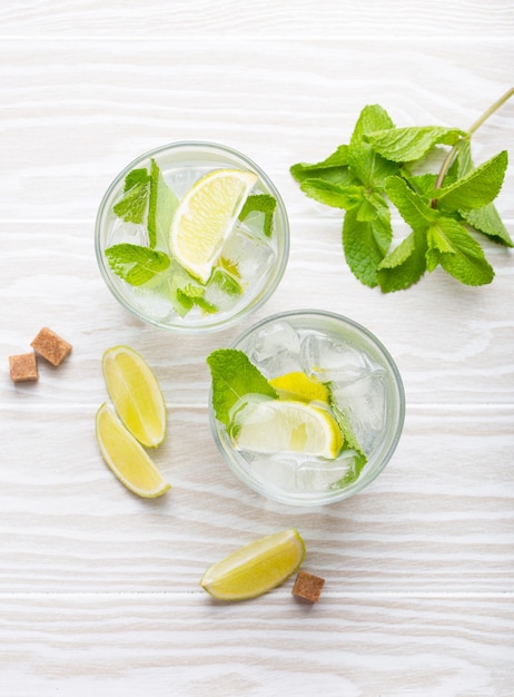 Bevande estive con ghiaccio, lime e menta fresca in bicchieri su fondo rustico in legno bianco. vista dall'alto di limonate/mojito rinfrescanti fredde, ideali per feste estive e divertimento, dall'alto, primo piano