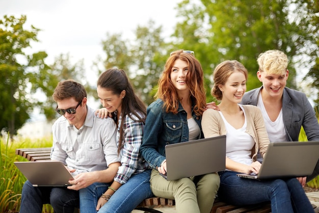 summer, education, technology and people concept - group of students or teenagers with laptop computers sitting on bench outdoors