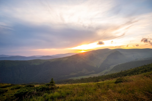 Summer evening mountain landscape with grassy hills and distant peaks at colorful sunset.