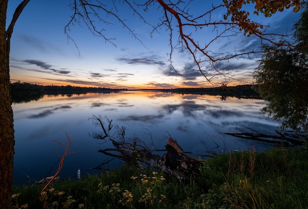 Summer evening twilight view on picturesque plain lake with forest and blossoming meadows on shores and sunset clouds reflections in water surface