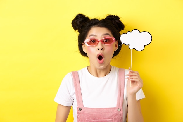 Summer and fashion concept. Excited party girl in sunglasses, holding comment cloud party mask and gasping amazed, standing impressed on yellow.