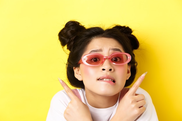 여름 패션 개념. 걱정 된 우유부단 한 아시아 소녀의 얼굴 만, 손가락을 옆으로 가리키고 두 가지 방법을 보여주는, 선글라스를 착용하고 선택할 수 없습니다.