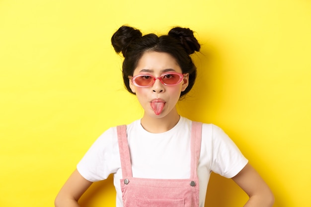 Photo summer and fashion concept. silly asian teen girl showing tongue, wearing sunglasses, standing on yellow.