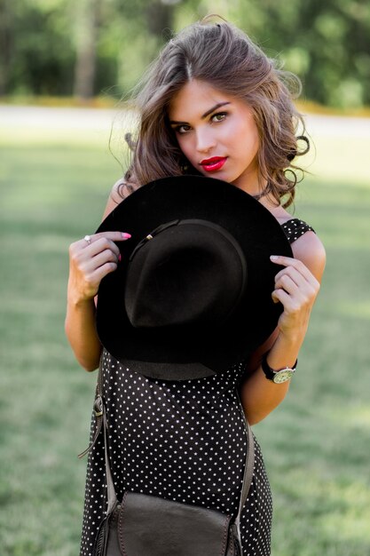 Summer fashion portrait of elegant woman with perfect wavy hair  in stylish elegant black hat and bright make up posing in the park. Street style.