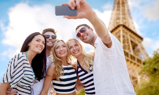 夏、フランス、観光、テクノロジー、人々のコンセプト – パリの背景にあるエッフェル塔の上で、スマートフォンで自撮りをする笑顔の友人のグループ