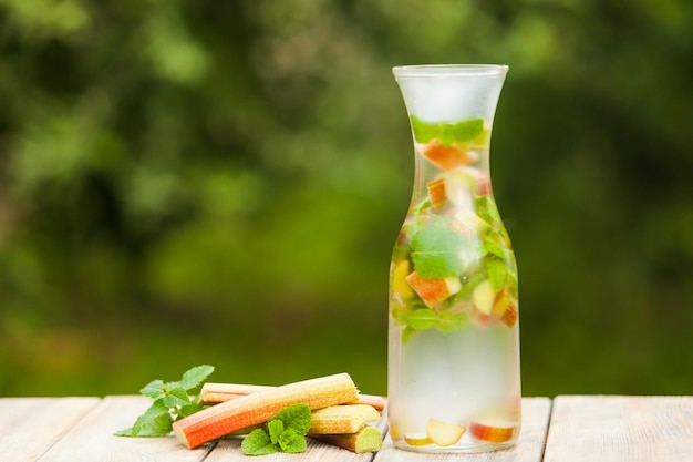 Summer fresh cold water with rhubarb and mint in a glass, the stems pieplant on the background on wooden table