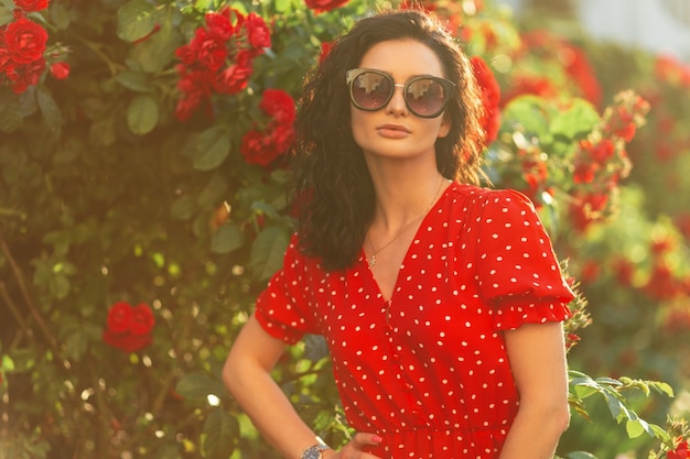 Summer fresh portrait of a beautiful young woman with fashionable sunglasses in a stylish red dress stands near a bush with flowers of roses in the garden